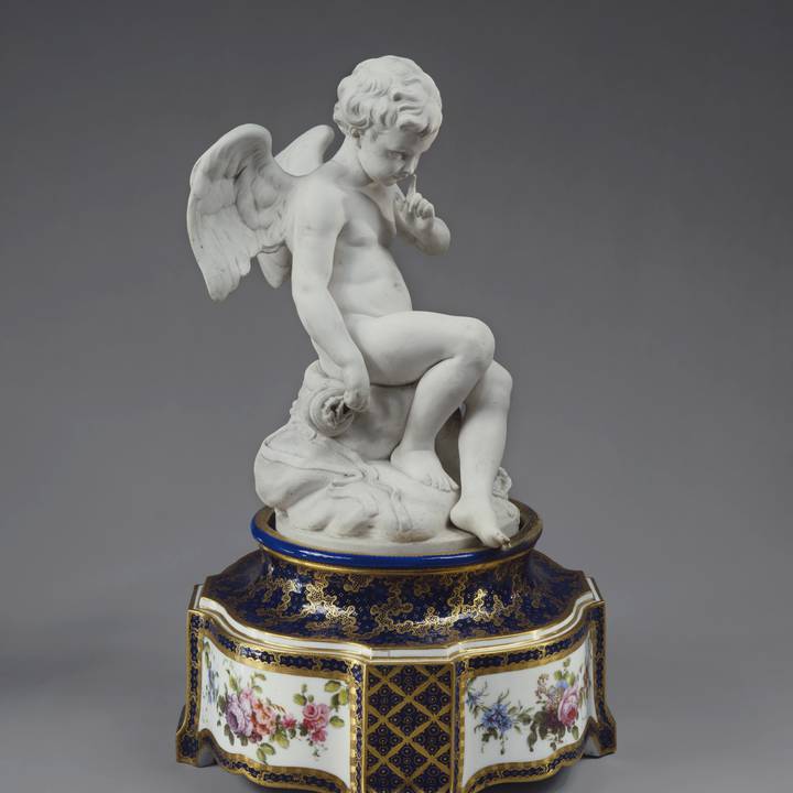 Porcelain sculpture of cupid sat on floral painted pedestal