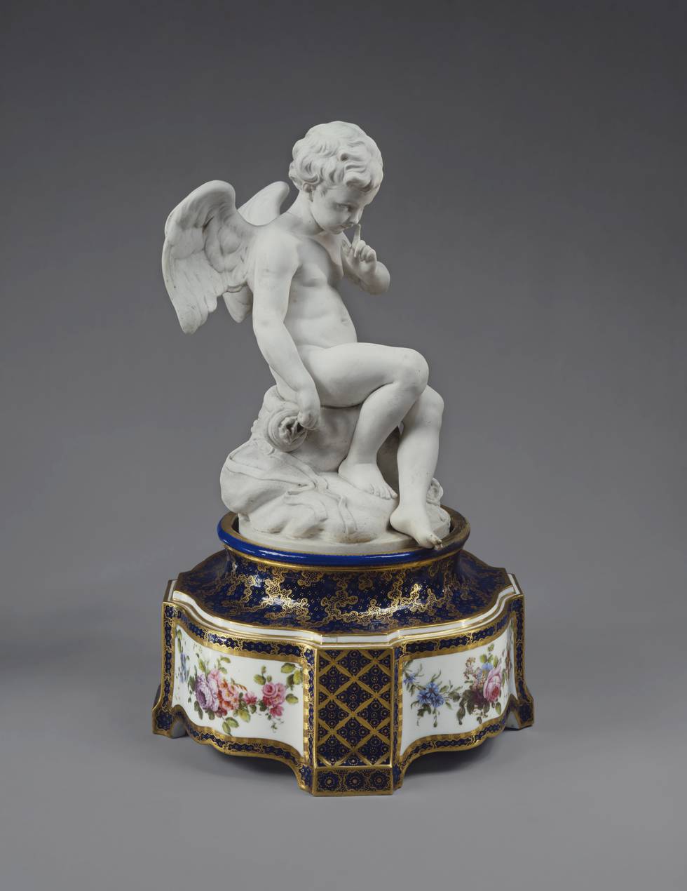 Porcelain sculpture of cupid sat on floral painted pedestal